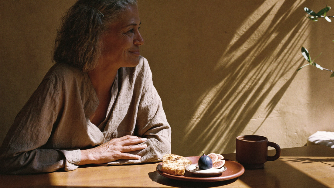 Woman in her 50s with silver hair enjoying a calm breakfast of figs and toast in warm morning light
