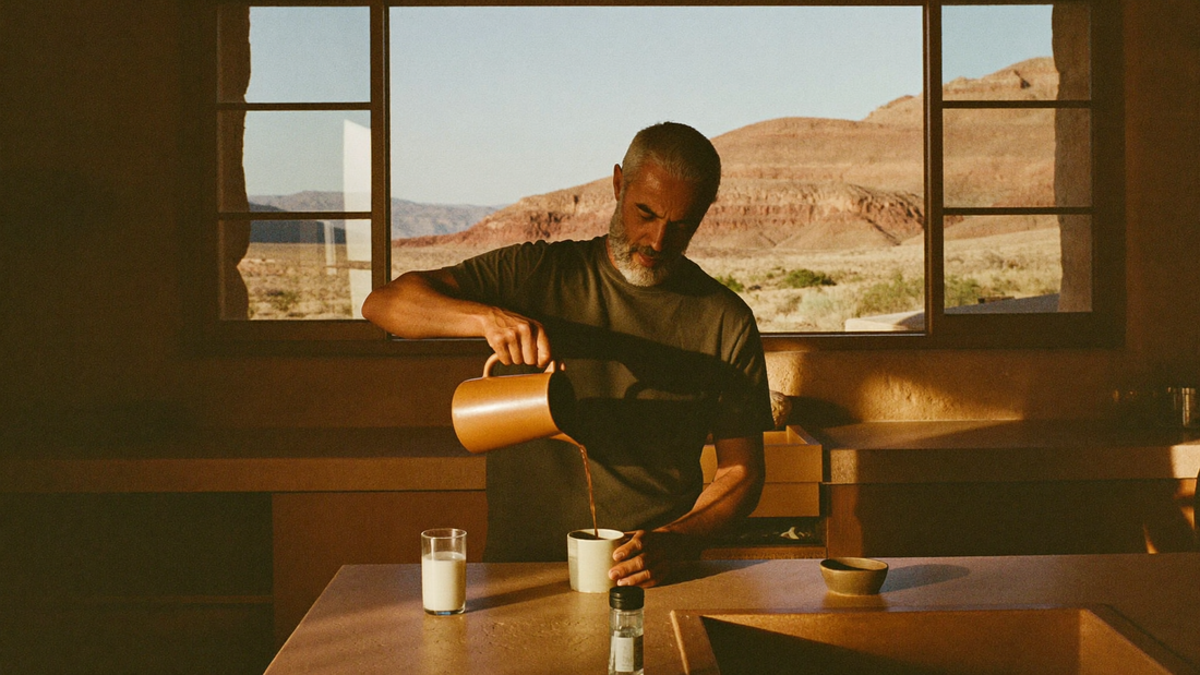 Man in his 60s with a grey beard pouring morning coffee at a stone kitchen counter with desert hills outside