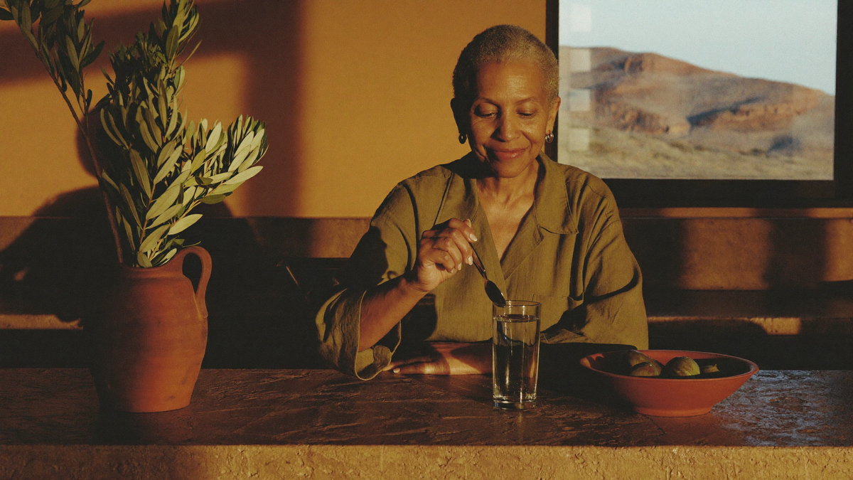 Woman in her late 50s stirring a glass of water at a sandstone kitchen counter in warm morning light
